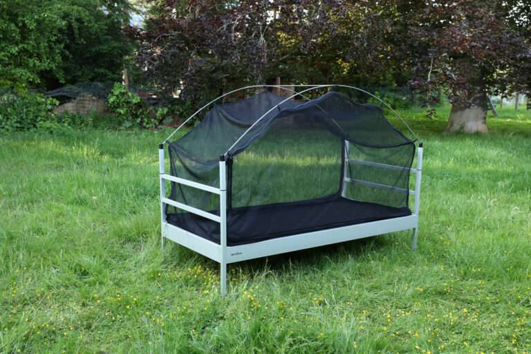 Die SkyHeia, das Outdoor-Bett zum Draußen Schlafen, steht auf einer Wiese. Es ist weder Matratze noch Bettwäsche aufgelegt. Das Bett ist jedoch umhüllt von einem schwarzen Moskitonetz.