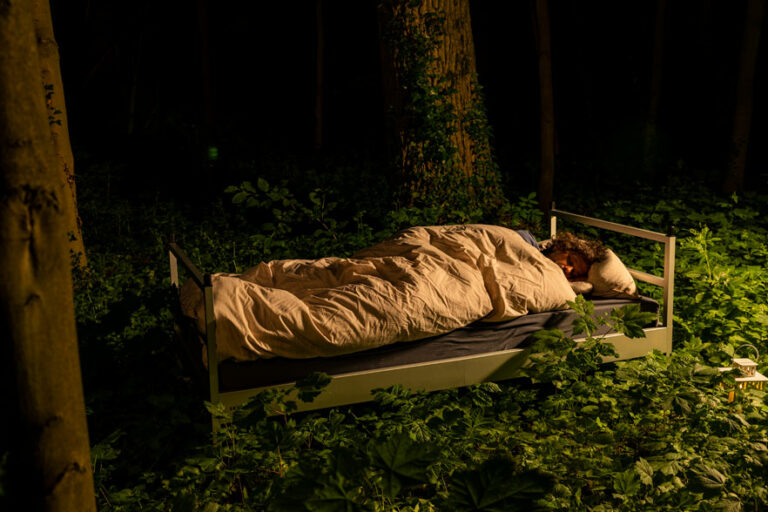 Draußen Schlafen unter freiem Himmel macht glücklick: Das Bild zeigt ein Bett, welches im Wald steht und in dem jemand mit glücklichem Gesichtsausdruck schläft