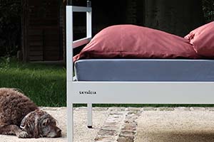 Die Hündin Laika liegt gemütlich neben dem Outdoor Bett SkyHeia. Das Besondere am Bett ist: es ist gemütlich wie ein richtiges Bett. Das Spannbettlaken ist grau, das Kissen ist Bordeaux-Rot.